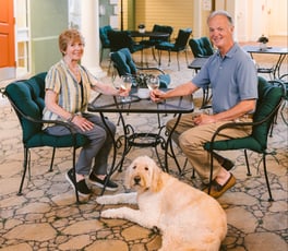 Two residents enjoy time together with their dog in a bright, welcoming indoor commons area. Seated at a café-style table with drinks, they relax in a warm, pet-friendly setting designed to encourage connection, comfort, and everyday enjoyment.