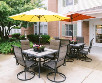 An inviting outdoor courtyard features comfortable seating, colorful umbrellas, and beautifully landscaped gardens. This peaceful space offers residents a welcoming setting to relax, socialize, enjoy fresh air, and connect with nature in a safe, serene environment.