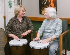 Two women share a joyful moment during a drumming activity, smiling and engaging with one another. The image highlights the power of music, connection, and meaningful social interaction in creating a vibrant and enriching community experience.