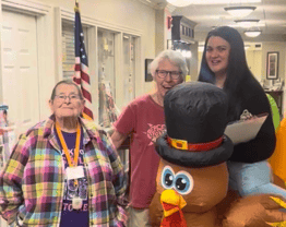 Three people gather in a community space beside a festive turkey decoration, smiling for the camera. The image captures a fun seasonal celebration that brings residents and team members together through shared activities and community spirit.