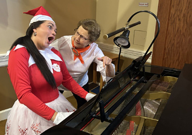 Two women share a joyful musical moment at a piano, one dressed in a festive holiday costume while the other smiles beside her. The image captures the fun, laughter, and meaningful connections created through music and seasonal celebrations.
